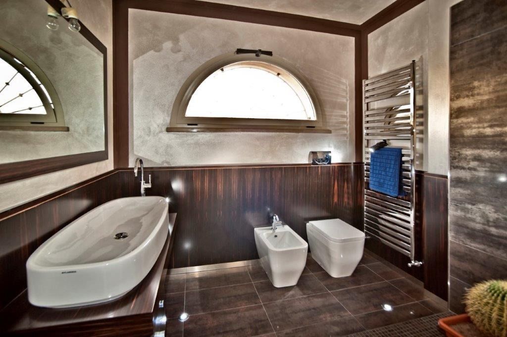 Bagno moderno con vasca freestanding, due WC bianchi, specchi e portasciugamani su pareti in legno scuro.