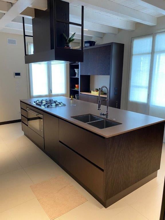 Cucina moderna con isola in legno scuro, lavello, piano cottura e ampie finestre con tende.